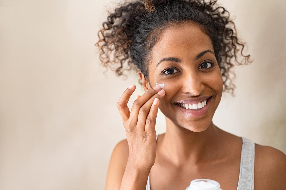 Smiling african girl with curly hair applying facial moisturizer while holding jar and looking at camera. Portrait of young black woman applying cream on her face isolated on beige background. Close up of happy attractive beauty woman caring of her skin standing on light brown wall with copy space.