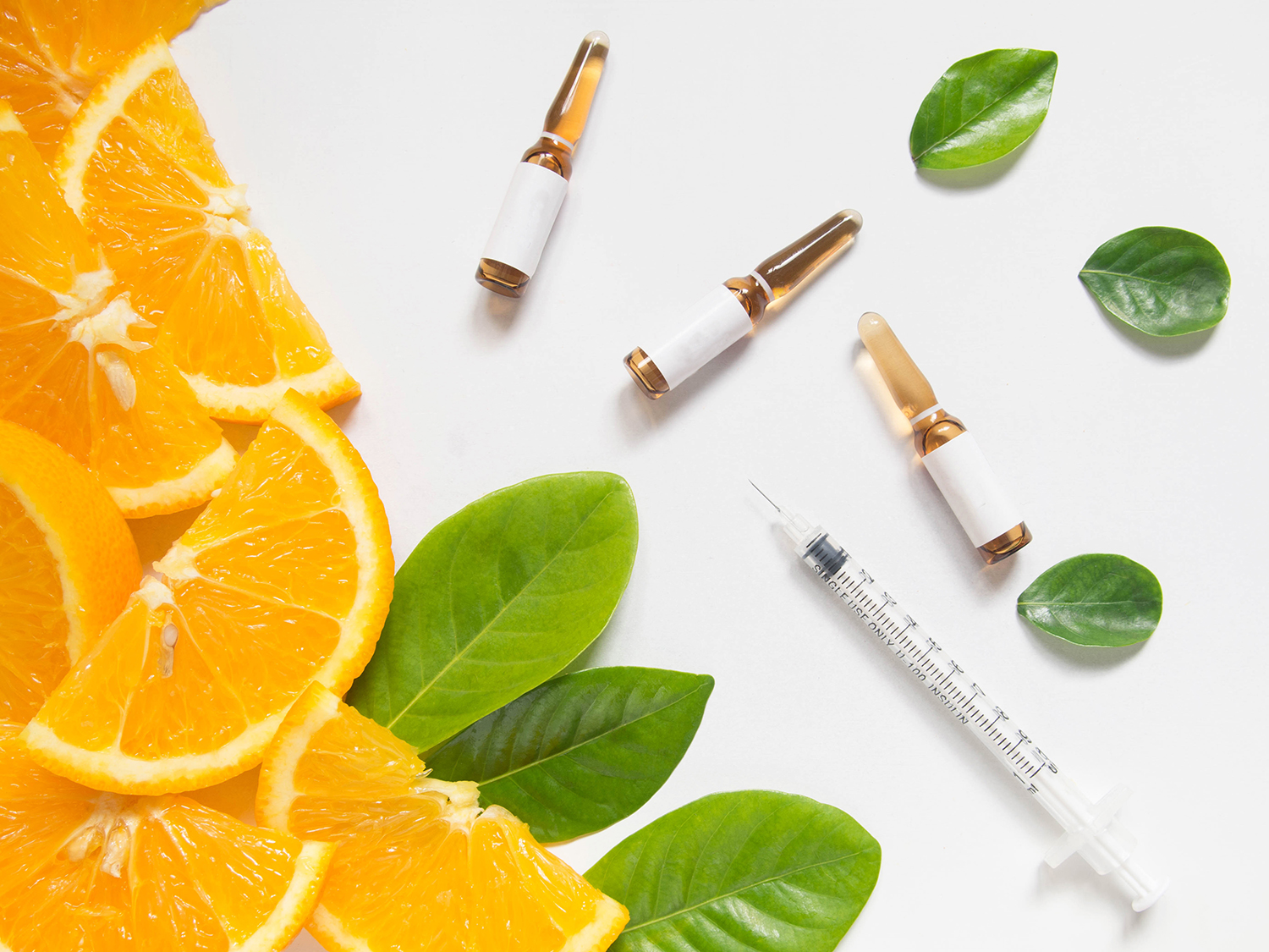 Top view of vitamin C brown ampule for injection and syringe with fresh juicy orange fruit slice and green leaves on white table. Vitamin/mineral supplement concept. Beauty product mock-up.
